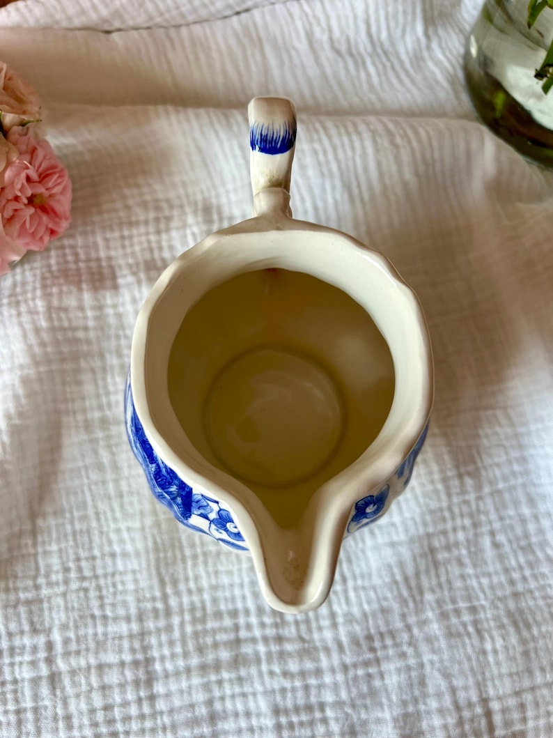 May include: Cream-colored ceramic creamer with blue floral patterns and a spout. The handle is accented with blue paint. The creamer is empty and sits on a white, textured surface.