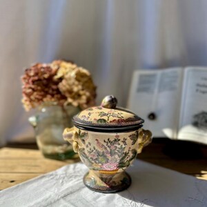 Peut inclure: Un pot en céramique avec couvercle, présentant une base de couleur crème ornée d'un motif polychrome détaillé d'oiseaux, de fleurs et de feuillage. Le récipient possède deux anses latérales, une base sur pied et un petit bouton sur le couvercle.