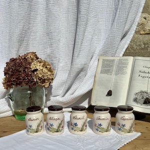Könnte beinhalten: Fünf kleine Gewürzdosen aus Keramik mit dunkelbraunen Deckeln, jeweils mit handgemalten Blumenmotiven und den französischen Beschriftungen Laurier, Estragon, Fenouil, Romarin und Paprika. Die Dosen stehen in einer Reihe auf einem weißen Tuch.