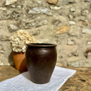 Peut inclure: Un pot en céramique brun foncé de forme cylindrique avec un bord arrondi et une base légèrement effilée. Le récipient présente une texture mate et patinée sur toute sa surface extérieure, affichant une teinte terreuse profonde.