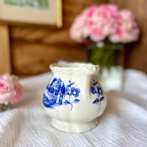 May include: A small, white ceramic vase with a blue floral design. The vase has a scalloped rim and a bulbous shape, sitting on a white cloth. The background includes a blurred image and pink flowers.