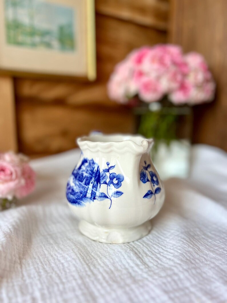 May include: A small, white ceramic vase with a blue floral design. The vase has a scalloped rim and a bulbous shape, sitting on a white cloth. The background includes a blurred image and pink flowers.