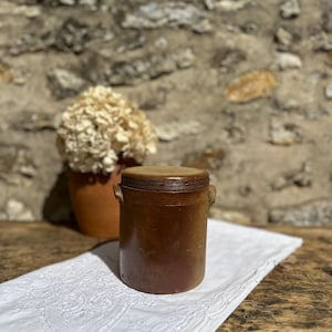 May include: A cylindrical brown stoneware crock with a flat lid and two small side handles. The vessel features a smooth, dark glazed finish with subtle horizontal ridges around the upper rim, presenting a sturdy and functional kitchen storage container.