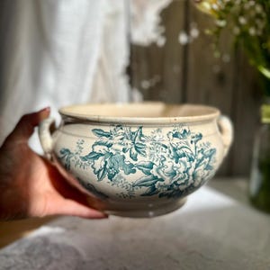 May include: A cream-colored ceramic bowl featuring a dark teal floral transferware pattern. The bowl has a rounded body, a wide opening, and two small side handles. The surface displays visible fine crackling throughout the glazed finish.