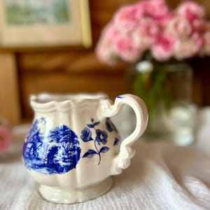 May include: Cream-colored ceramic pitcher with blue floral and landscape designs. The pitcher has a scalloped rim and a decorative handle. A bouquet of pink flowers is in the background.