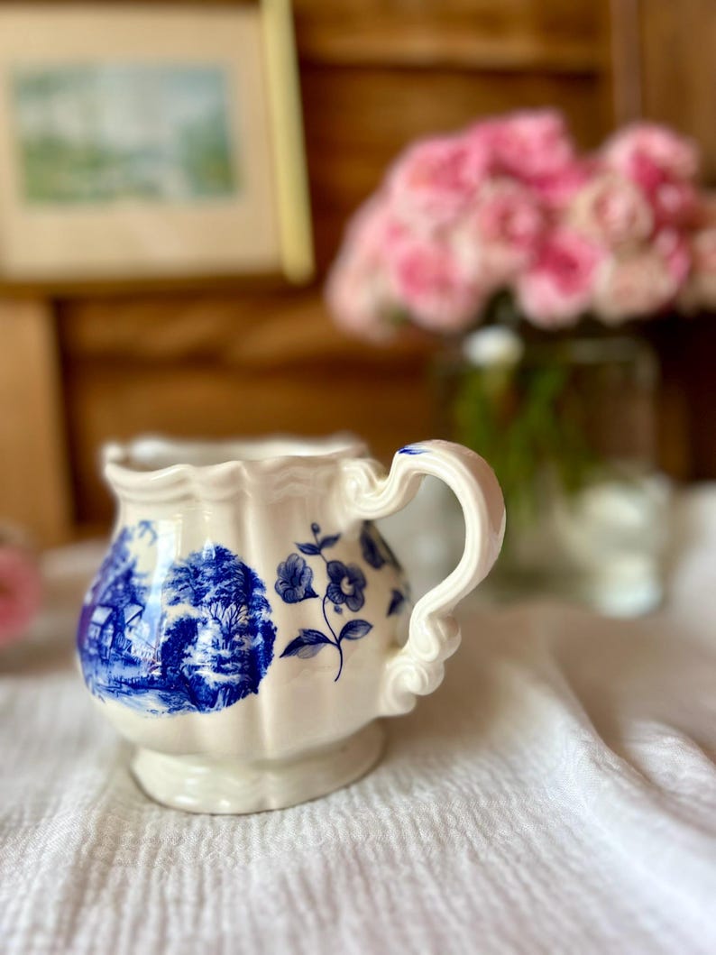 May include: Cream-colored ceramic pitcher with blue floral and landscape designs. The pitcher has a scalloped rim and a decorative handle. A bouquet of pink flowers is in the background.