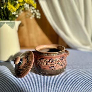 Op de afbeelding: Een kleine, open keramische suikerpot met een deksel. De kom is roze met een bloemenmotief en twee kleine handvatten. Het deksel ligt ernaast. De kom staat op een blauw-wit gestreept tafelkleed.