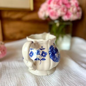 May include: Cream-colored ceramic creamer with blue floral and landscape designs. The creamer has a handle and a scalloped rim. The design features blue flowers and a rural scene. The creamer is sitting on a white cloth.