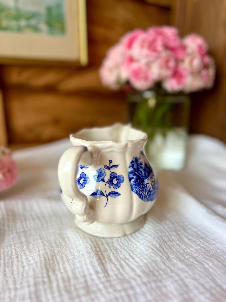 May include: Cream-colored ceramic creamer with blue floral and landscape designs. The creamer has a handle and a scalloped rim. The design features blue flowers and a rural scene. The creamer is sitting on a white cloth.