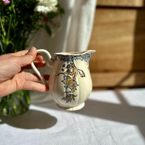 Peut inclure: Pichet en céramique crème avec une anse incurvée et un bec verseur. Il est orné de motifs floraux bleus et dorés. Le pichet est tenu par une main, avec un bouquet de fleurs en arrière-plan.