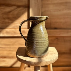 May include: A ceramic pitcher with a glossy olive green glaze and a textured, ribbed body. The vessel features a curved handle on the side, a wide mouth with a small pouring spout, and a flat, unglazed base showing the natural clay color.