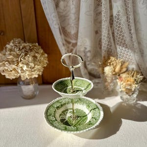 May include: A two-tiered serving stand featuring green and white transferware plates with a detailed pastoral landscape pattern. The stand has a central polished silver-tone metal rod and a matching oval handle at the top for carrying.