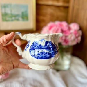 May include: A white porcelain creamer with a blue landscape design. The creamer has a decorative handle and a scalloped rim. The blue design depicts a scenic landscape. The creamer is a vintage piece, perfect for serving cream or milk.