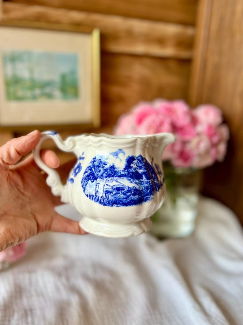 May include: A white porcelain creamer with a blue landscape design. The creamer has a decorative handle and a scalloped rim. The blue design depicts a scenic landscape. The creamer is a vintage piece, perfect for serving cream or milk.