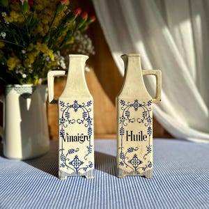 以下が含まれることがあります： 取っ手付きの2つのオフホワイトのセラミッククルエットボトル。青い花の模様で装飾されています。1つのボトルには「Vinaigre」、もう1つには「Huile」と書かれています。ボトルは青と白のストライプのテーブルクロスの上にあります。