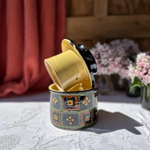 May include: A ceramic butter dish with a lid, featuring a yellow interior and a black rim. The exterior is decorated with a repeating pattern of squares in black, orange, and green. The dish is sitting on a white tablecloth.
