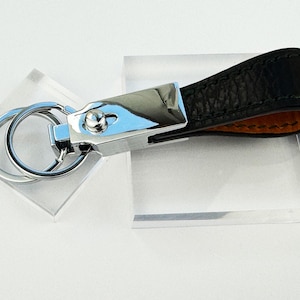 May include: A black leather keychain with a silver metal clasp and key rings. The leather strap has a textured surface and a contrasting orange interior. The keychain is displayed on a clear acrylic block.