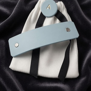 May include: A light blue rectangular leather cuff with two silver-toned metal rivets, attached to a white fabric drawstring pouch by a black ribbon. A circular light blue leather tag with the gold-colored letter A is centered at the top.