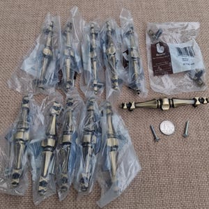 May include: A collection of ten antique brass-finished metal cabinet pulls, each featuring a central bulbous shape and tapered ends. Nine pulls are individually wrapped in clear plastic, while one is unwrapped alongside two metal screws and a US quarter.