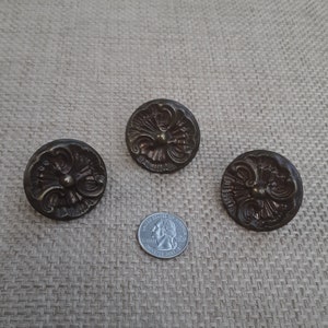 May include: Three antique brass drawer pulls with ornate floral designs. The pulls are round and have a raised center with a small knob. The pulls are about the size of a quarter.