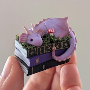 Puede incluir: Una figura de drag&oacute;n p&uacute;rpura en miniatura que descansa sobre una pila de tres libros. El drag&oacute;n tiene una cola larga, alas y cuernos. Una peque&ntilde;a seta roja y blanca est&aacute; cerca. Los libros son negros, morados y blancos.