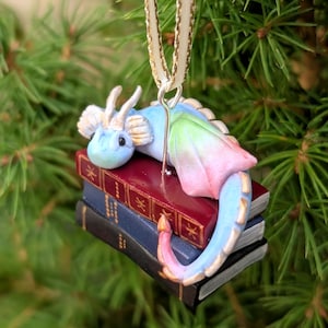 May include: A fantasy-themed Christmas tree ornament. A small, colourful dragon with blue, pink, and green hues rests on a stack of miniature books. The ornament is suspended by a cream ribbon, ideal for festive decoration.
