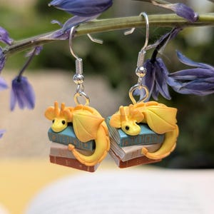 Two earrings dangle from a horizontal stem of bluebells. There are three books, rust, tan and sage, with gold detail on the front. On top of them sit a golden yellow dragon, with their tail wrapped around the front of the books.