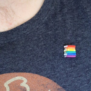 Könnte beinhalten: Ein dunkelgraues T-Shirt mit einem verblassten orangefarbenen und weißen Aufdruck, an dem ein Stapel kleiner Regenbogenbücher befestigt ist.