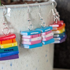 May include: Three pairs of earrings with a book theme. Each earring features a stack of colourful books with silver-tone fish hook ear wires. The books are in rainbow colours, with some featuring the transgender flag colours.