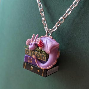 A tiny pendant of a purple dragon with gold detail sits on three books, green, purple and black, which have gold detail on their spines. There is moss around the dragon and a tiny red and white spotted mushroom. It hangs from a silver chain against a dark green background.