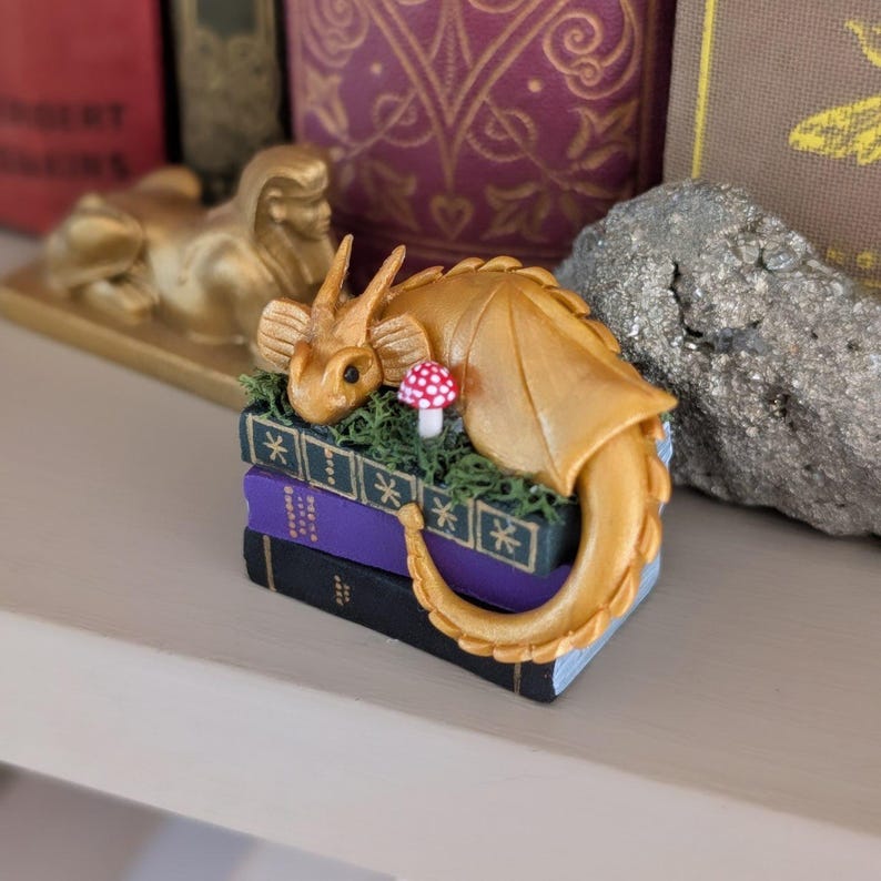 Puede incluir: Una figura de drag&oacute;n dorado descansando sobre una pila de libros en miniatura. El drag&oacute;n tiene alas detalladas y est&aacute; rodeado de musgo verde y una peque&ntilde;a seta con lunares rojos y blancos. Los libros son morados y negros.