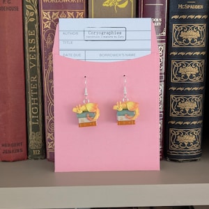 May include: Dangle earrings with a yellow and orange dragon design sitting on books. The earrings are presented on a pink card with the text "Coryographies Handmade Creations by Cory". The background shows a shelf with antique books.