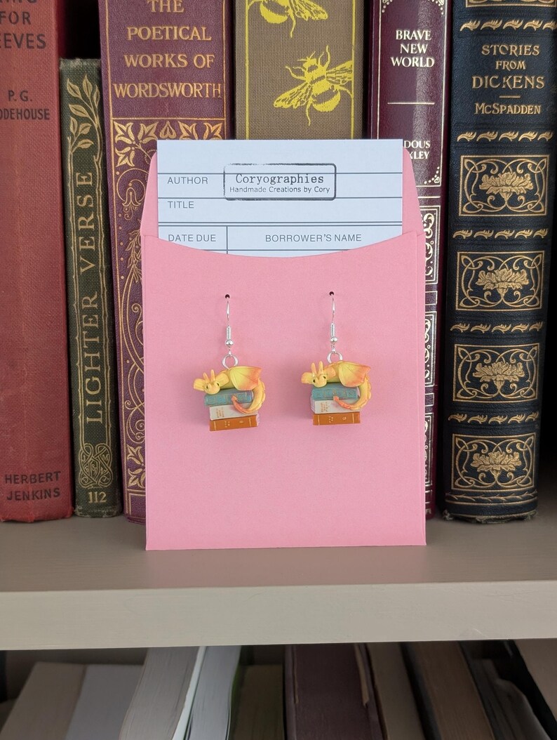 May include: Dangle earrings with a yellow and orange dragon design sitting on books. The earrings are presented on a pink card with the text "Coryographies Handmade Creations by Cory". The background shows a shelf with antique books.