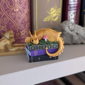Puede incluir: Una figura de drag&oacute;n dorado que descansa sobre una pila de libros en miniatura. El drag&oacute;n tiene alas detalladas y una cola enroscada. Tambi&eacute;n hay un peque&ntilde;o hongo rojo y blanco. Los libros son morados y negros.