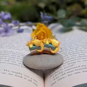 May include: Earrings with a fantasy theme, each featuring a yellow dragon on a stack of books. The dragon has blue details. The earrings are set on a grey stone, with a book and flowers in the background.
