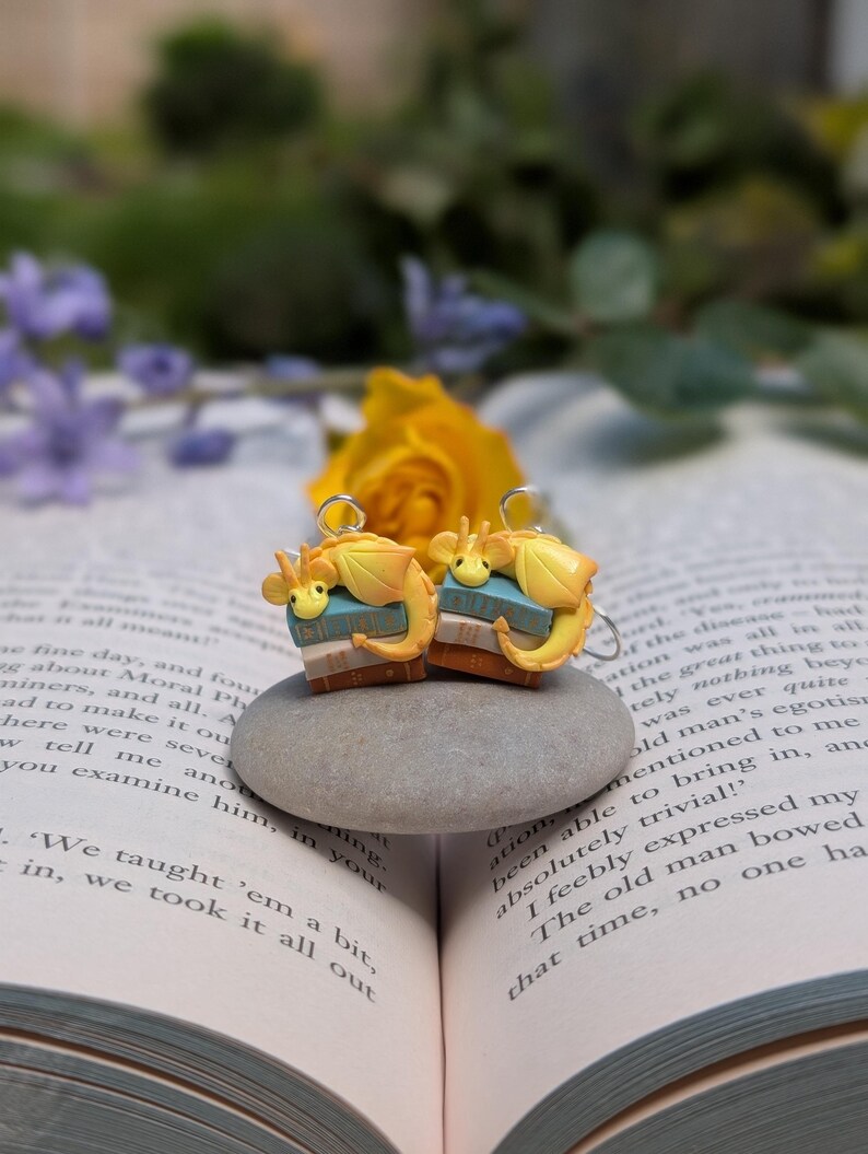May include: Earrings with a fantasy theme, each featuring a yellow dragon on a stack of books. The dragon has blue details. The earrings are set on a grey stone, with a book and flowers in the background.