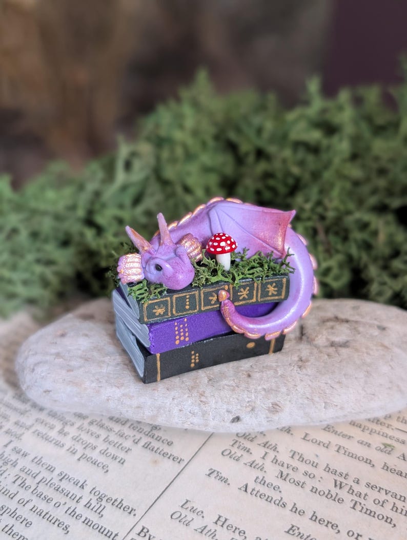 Puede incluir: Una figura en miniatura de un drag&oacute;n p&uacute;rpura descansando sobre una pila de tres libros. El drag&oacute;n tiene detalles dorados y un hongo rojo y blanco. Los libros son negros y morados con detalles dorados.