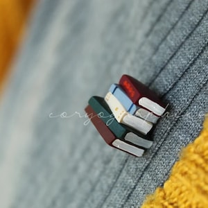 May include: A stack of miniature books with red, blue, green, and white covers. The books are made of polymer clay and are a cute and whimsical accessory.