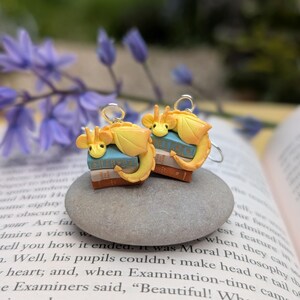 May include: Two charming earrings, each with a yellow dragon design atop a tiny stack of books. The dragons have orange detailing and are suspended from silver-coloured hooks. The earrings are presented on a grey stone, with a blurred backdrop of a book and flowers.