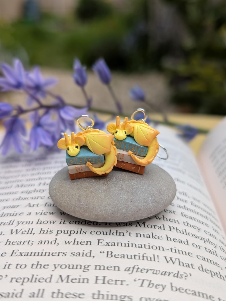 May include: Two charming earrings, each with a yellow dragon design atop a tiny stack of books. The dragons have orange detailing and are suspended from silver-coloured hooks. The earrings are presented on a grey stone, with a blurred backdrop of a book and flowers.