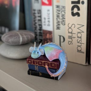 May include: A small, blue and pastel-colored dragon figurine resting on a stack of miniature books. The dragon has a light blue body, with pink and green accents on its wings and tail. The books are burgundy, blue, and black.