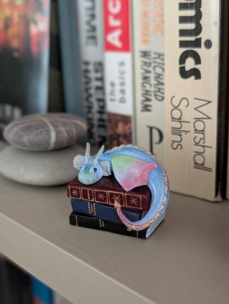 May include: A small, blue and pastel-colored dragon figurine resting on a stack of miniature books. The dragon has a light blue body, with pink and green accents on its wings and tail. The books are burgundy, blue, and black.