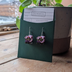 May include: A pair of dangle earrings featuring a purple dragon perched atop a stack of miniature books. The earrings are displayed on a dark green card with a white label that reads "Coryographies Handmade Creations by Cory". The background includes a plant in a pot.