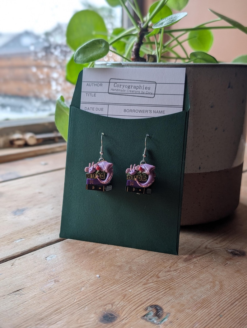 May include: A pair of dangle earrings featuring a purple dragon perched atop a stack of miniature books. The earrings are displayed on a dark green card with a white label that reads "Coryographies Handmade Creations by Cory". The background includes a plant in a pot.