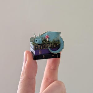 Puede incluir: Una figura de drag&oacute;n azul en miniatura que descansa sobre una pila de tres libros. El drag&oacute;n tiene una cola enroscada y est&aacute; rodeado de follaje verde y una seta roja y blanca. Los libros son negros y morados.