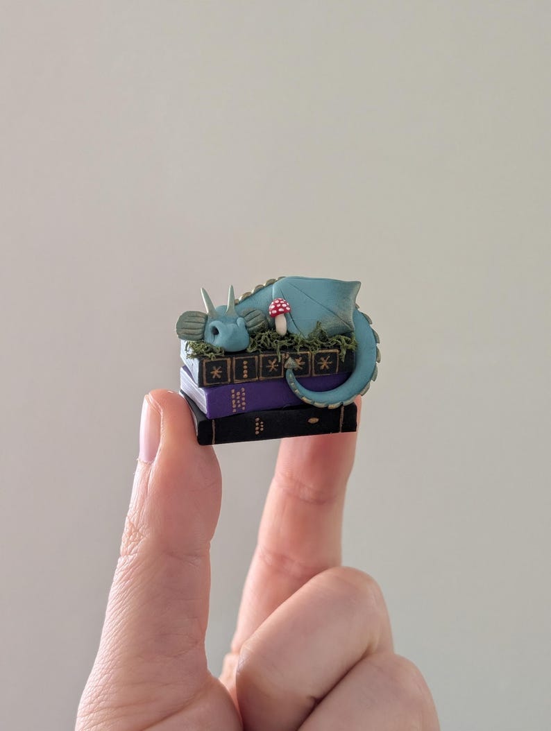 Puede incluir: Una figura de drag&oacute;n azul en miniatura que descansa sobre una pila de tres libros. El drag&oacute;n tiene una cola enroscada y est&aacute; rodeado de follaje verde y una seta roja y blanca. Los libros son negros y morados.