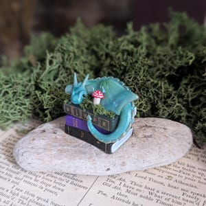 Puede incluir: Una escena de fantas&iacute;a en miniatura con una figura de drag&oacute;n verde azulado descansando sobre una pila de libros. Una peque&ntilde;a seta roja y blanca est&aacute; cerca. El drag&oacute;n est&aacute; sobre una piedra, rodeado de musgo verde.