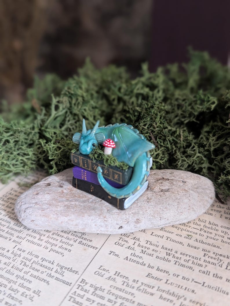 Puede incluir: Una escena de fantas&iacute;a en miniatura con una figura de drag&oacute;n verde azulado descansando sobre una pila de libros. Una peque&ntilde;a seta roja y blanca est&aacute; cerca. El drag&oacute;n est&aacute; sobre una piedra, rodeado de musgo verde.