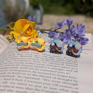 May include: Four pairs of dragon earrings. Each dragon sits atop a stack of books. The dragons are in yellow, blue, and pink hues. The earrings are presented on an open book, with yellow and purple flowers.