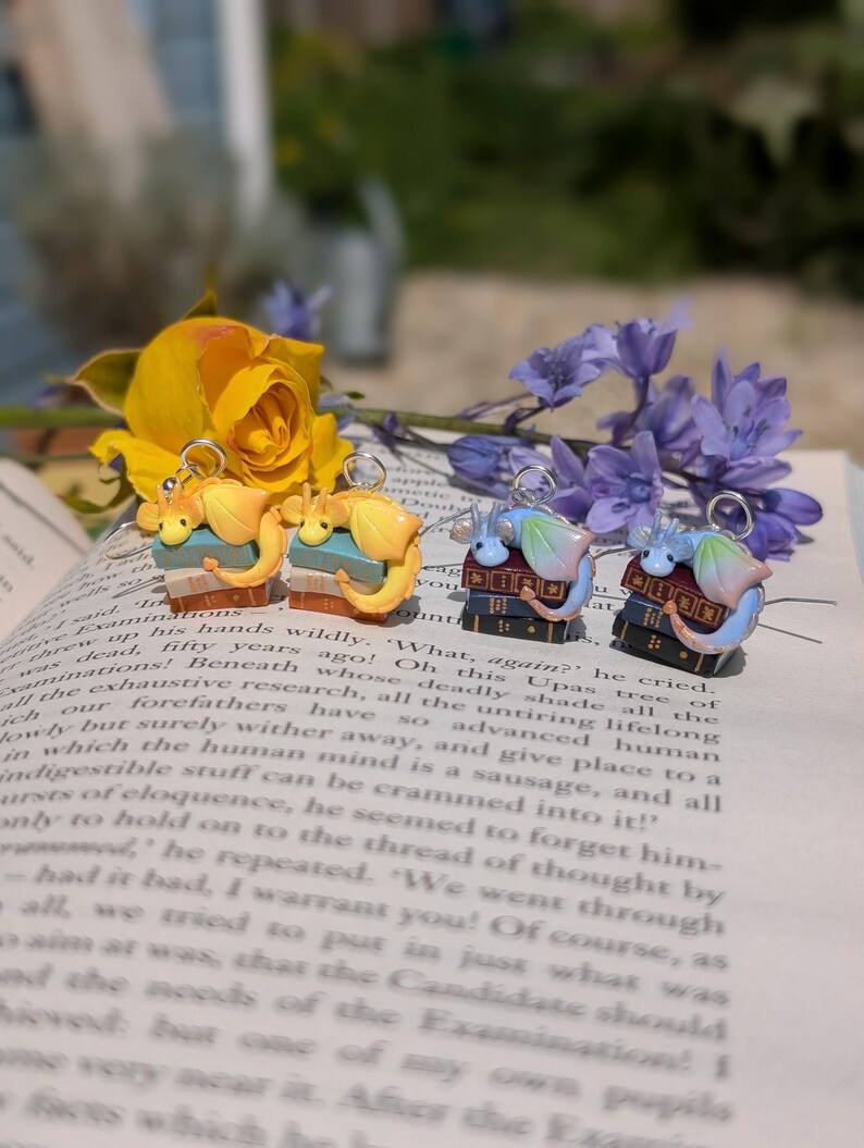 May include: Four pairs of dragon earrings. Each dragon sits atop a stack of books. The dragons are in yellow, blue, and pink hues. The earrings are presented on an open book, with yellow and purple flowers.